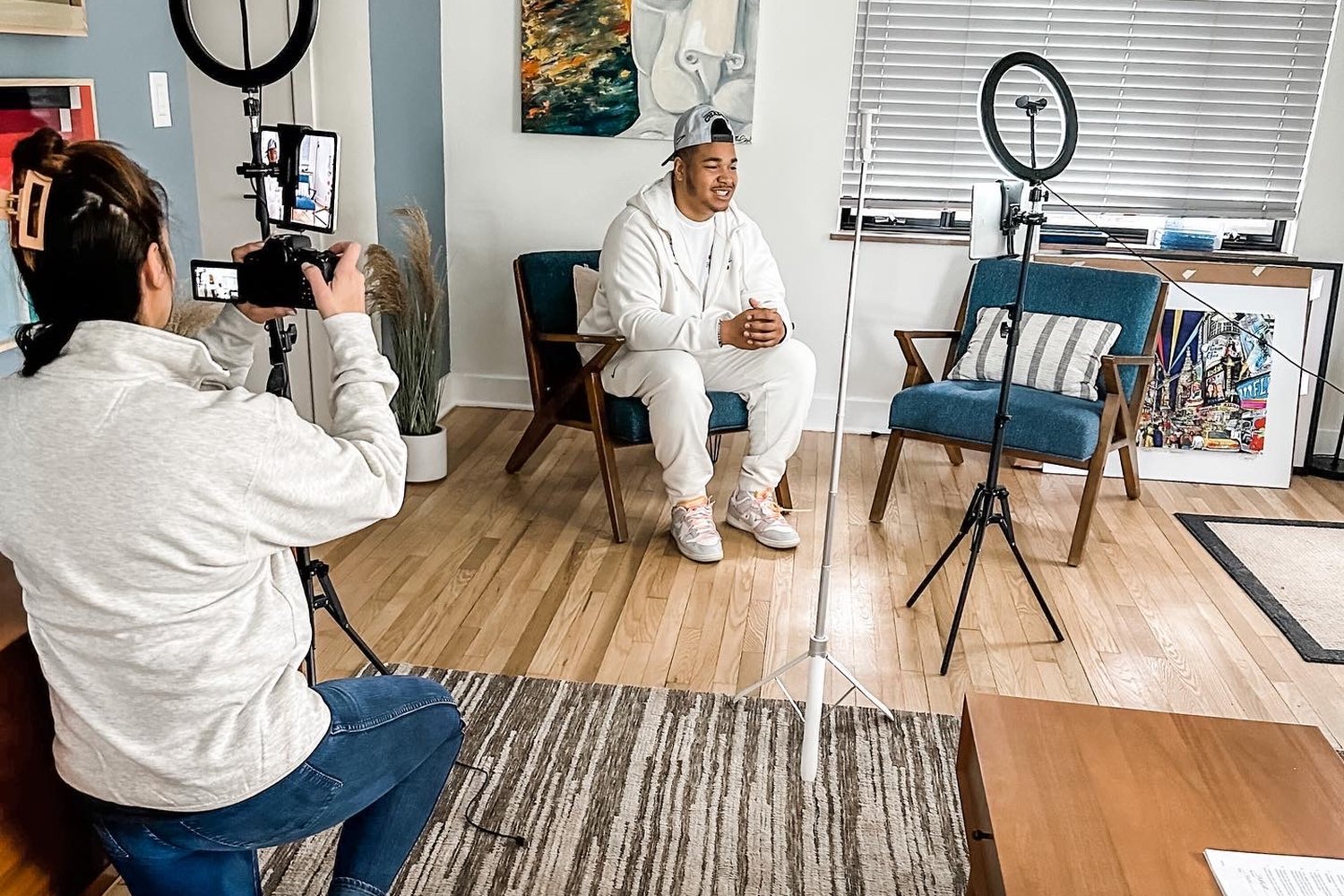 A shooter kneeling with a camera on a monitor rig, filming a college football player seated for an interview in a domestic location.