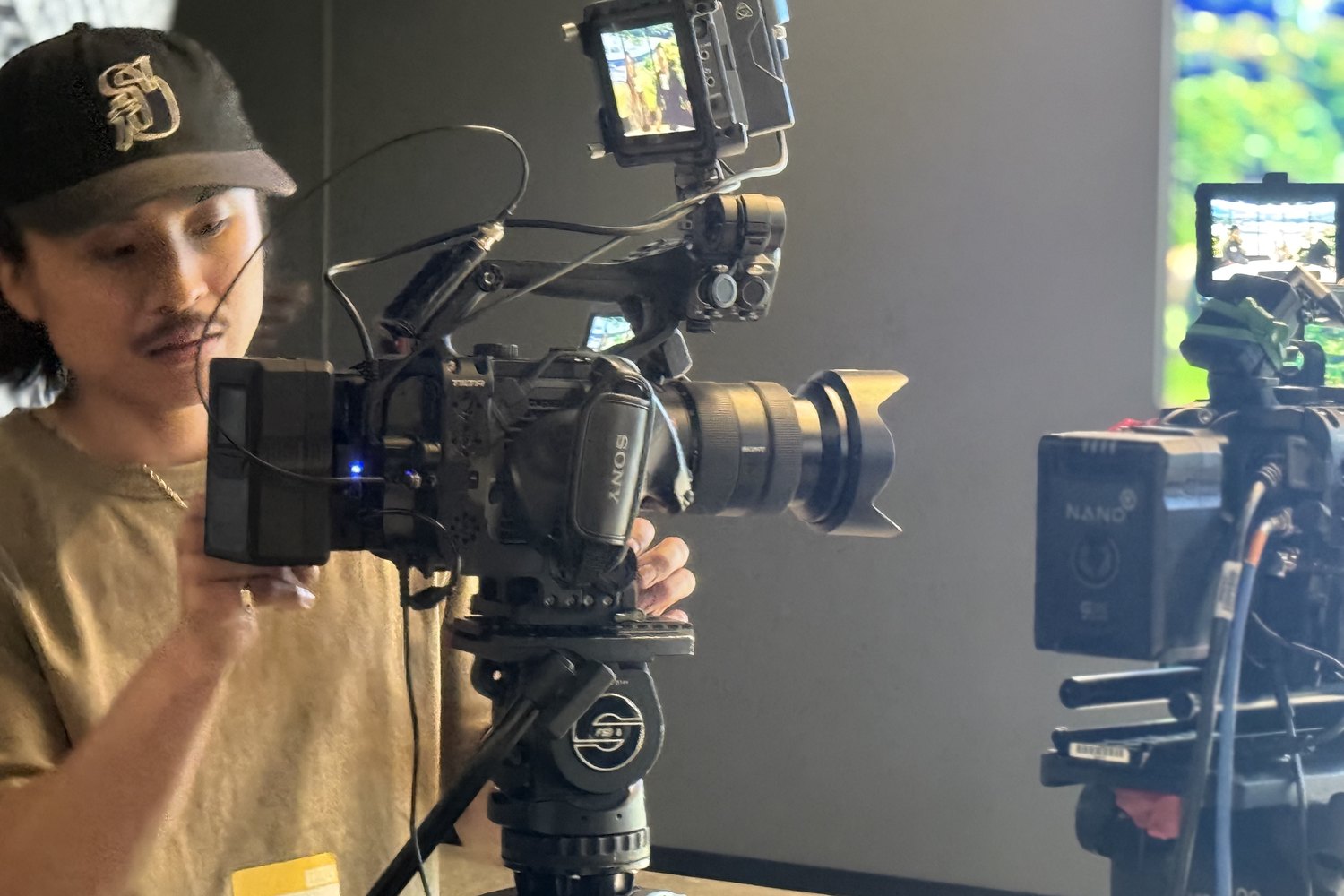 A camera operator in a cap steadying the A camera, a second rig and monitor beside him.