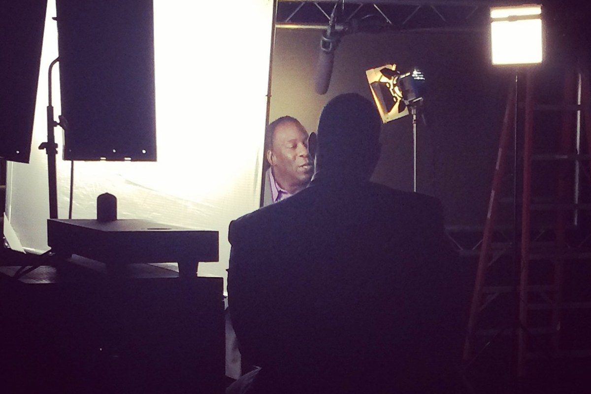Booker T lit against a bounce for a sit-down interview, the interviewer silhouetted in the foreground under a boom mic.
