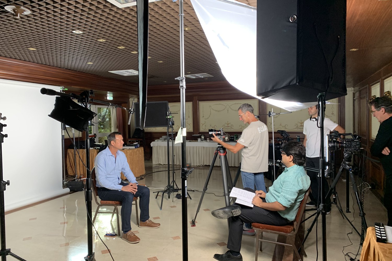 A multi-camera interview built inside a panelled room on location: large softbox overhead, A camera on sticks, a second camera on a slider, subject on one chair and the interviewer opposite with the run of questions.