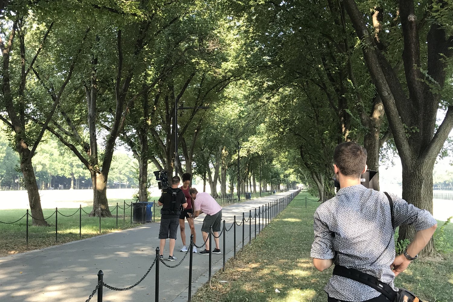 A location shoot with actors on a tree-lined path in Washington DC, camera operator working handheld in the shade.