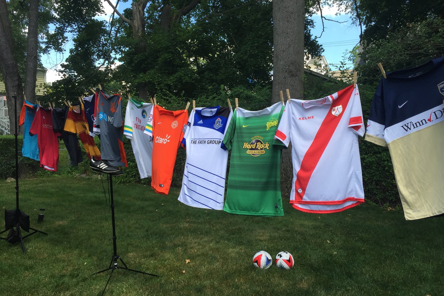 Club jerseys pegged along a line outdoors with two match balls on the grass, lit for a promo shoot.