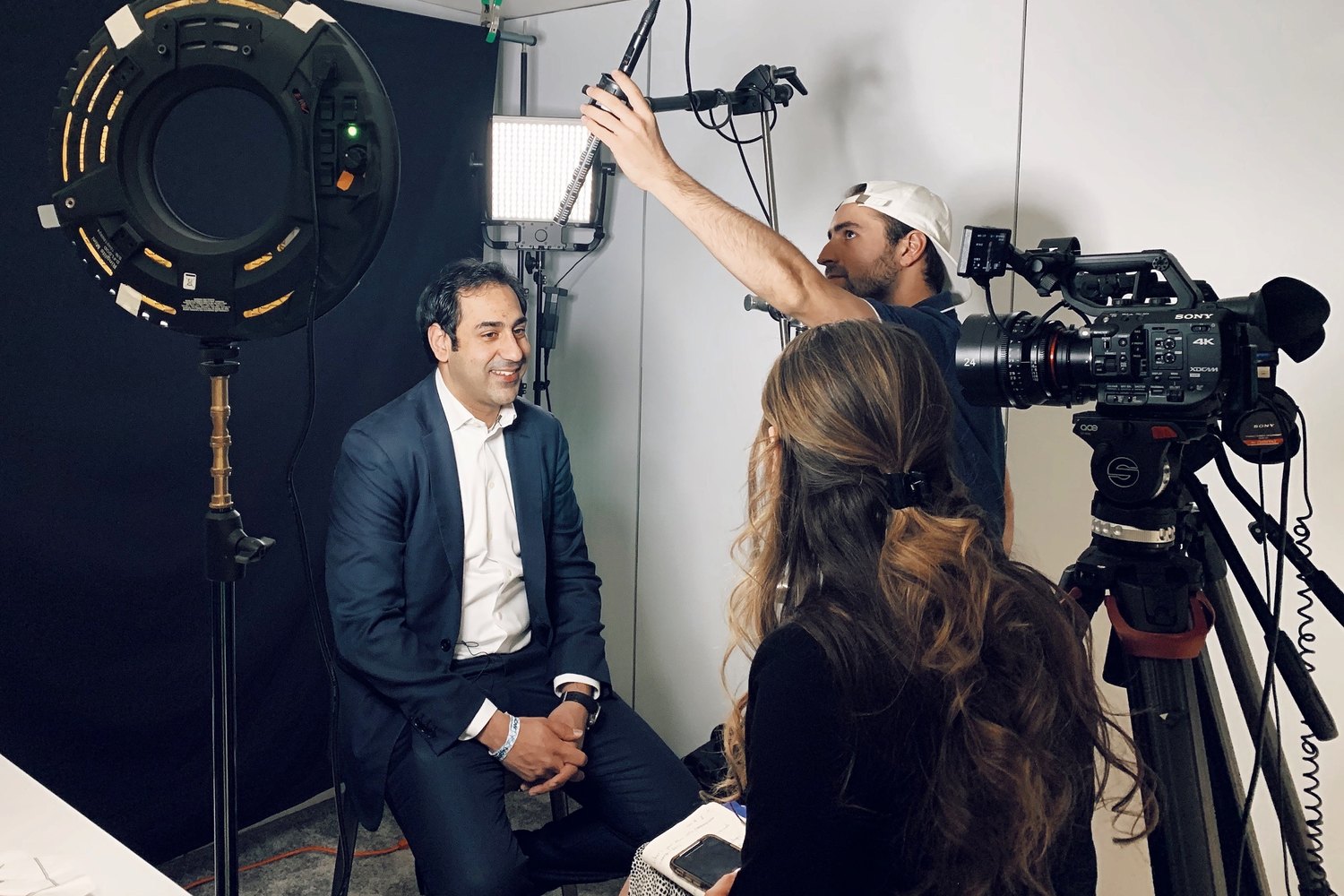A set built inside a plain meeting room on a conference site: ring light and panel up, boom operator reaching in overhead, subject in a suit ready to record.