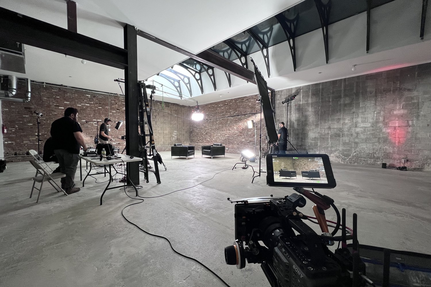 A raw brick loft rigged as a studio: skylight above, two chairs set for an interview, overhead frames flown, crew working the camera in the foreground.